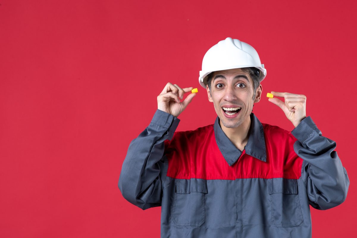 close up view curious young builder uniform with hard hat holding earplugs isolated red wall 3M Stock Soars on Military Assessment That Earplug Claimants Had No Hearing Loss